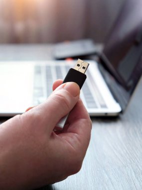 Bir adamın elindeki flaş belleği, tahta bir masada duran bir dizüstü bilgisayarın arka planına dayadığı dikey görüntü. Kavramsal görüntü. Seçici odak.