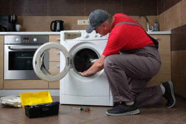 Üniformalı bir servis teknisyeni bir çamaşır makinesini tamir ediyor. Bir ev mutfağının arka planına karşı.