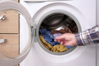 Bir adam plastik kapaktaki sıvı yumuşatıcıyı çamaşır makinesinin variline koyar. Sert musluk suyu koşullarında giysi bakımı.
