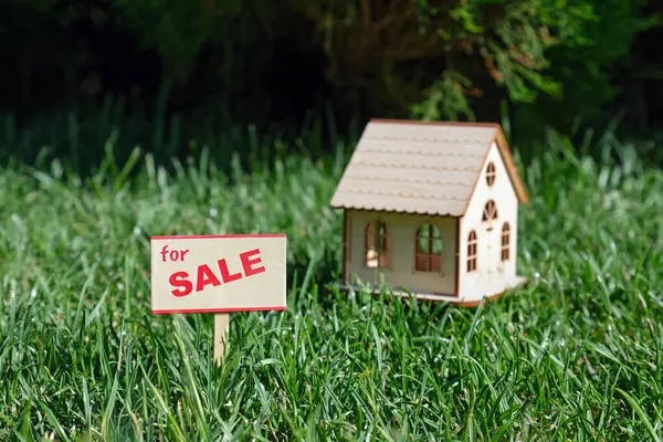 stock image Sign house for sale. A miniature composition with a toy wooden house in a meadow with green grass against a forest background. Conceptual image.