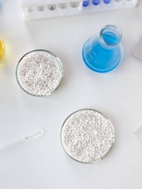 Kimyasal aletler ve cam eşyalar arka planında beyaz granülleri olan bir laboratuvar petri kabı. Dikey resim.