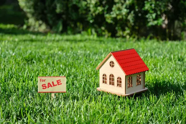 stock image Sign house for sale. A miniature composition with a toy wooden house with red rooftop in a meadow with green grass against a forest background. Conceptual image.