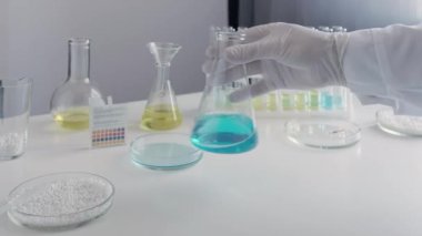 Bilimadamı mavi sıvıyı kimyasal bardakta karıştırıyor. Laboratuvar ekipmanlarının arka planına karşı.