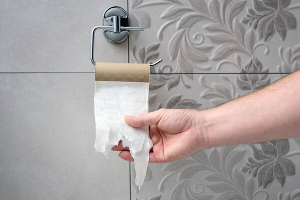 A man's hand reaches for an empty toilet paper holder on background tile wall. Shortage of paper.