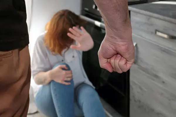 Aile içi şiddet sahnesi. Bir erkek yumruğuyla tehdit eder, ve bir kadın yerde oturur, kendini eliyle kapatır. Seçici odak.