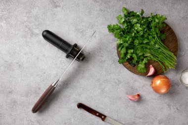 Close-up a kitchen knifes and hand sharpener on a table in a home kitchen. There is a cutting board, green coriander, onion, salt and garlic on the table. Preparing to cook.  clipart