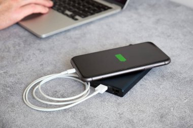 Bilgisayarın yanındaki masadaki enerji bankasından şarj olan modern bir siyah akıllı telefon. Tam pil gösterge aygıtı pili.