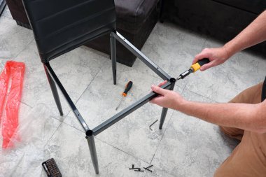 Kafkasyalı bir adam oturma odasında manuel tornavidayla sandalye yapıyor. Yeni bir dairede mobilya montajı. Yakın çekim, seçici odaklanma.