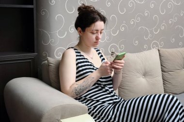 Kanepede oturan ev elbiseli, akıllı telefonlu ve üzgün yüzlü genç bir kadın. Kötü haber ya da mesajlar.