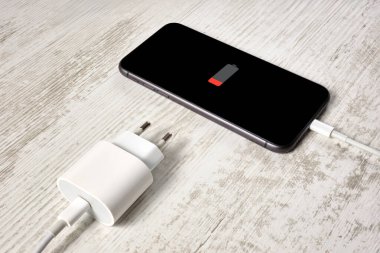 Düşük pil seviyeli modern bir akıllı telefon güzel bir vintage masanın üzerinde yatar. Cihaz kabloyla şarj cihazına bağlı. Kavramsal görüntü. Seçici odak.