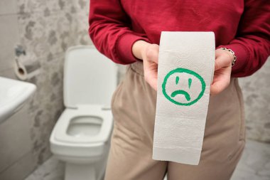 Tuvaletin yanında, üzerinde gülen surat çizilmiş tuvalet kağıdı tutan genç bir kadın. Tuvaletin önüne bir rulo tuvalet kağıdı koy..