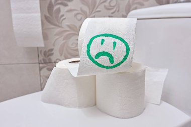 Üzücü, gülen yüzlü, yeşil renkli birkaç rulo tuvalet kağıdı dairenin banyosunun kapağında duruyor. Bağırsak ve sindirim sorununun kavramsal görüntüsü.