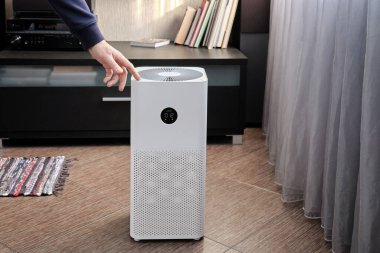 Bir adam pencerenin yanındaki oturma odasında büyük bir elektrikli hava temizleyiciyi çalıştırıyor. Büyük şehirlerdeki ekoloji ve hava temizliği sorunu.