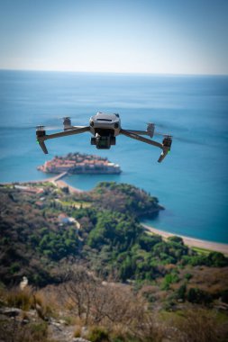 Arka planda bulanık bir kıyı manzarası ve adası olan dönen bıçaklarla uçan bir dronun yakın çekimine odaklanmış.. 