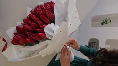 Beyaz kadın çiçekçi, lale buketinin dibinde kurdele bağlıyor. Bir profesyonel taze bir buket yapar. Küçük bir iş. Dikey video.