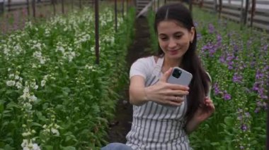 Çizgili önlüklü, gülümseyen ve poz veren bir kız akıllı telefonuyla matiola ile çiçek sıralarının arka planına selfie çekiyor. Ev yapımı ev yapımı sera. Aile işi ve iş