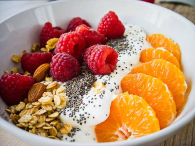 Kahvaltı masasında taze ahududu, mandalina dilimleri, granola ve chia çekirdeği ile kaplı canlı bir kase yoğurt.