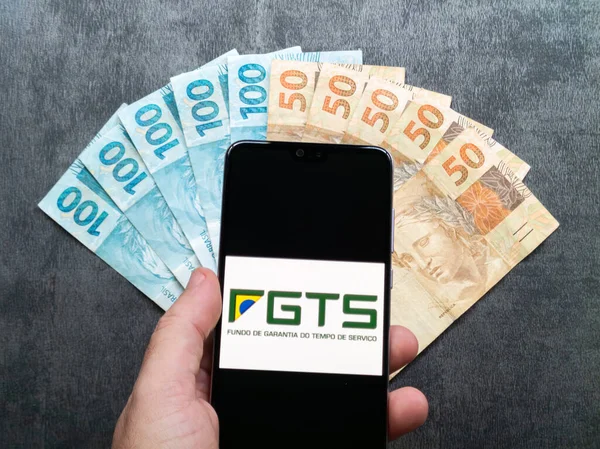 stock image Bahia, Brazil, 02 June 2023. Hand holding FGTS (Brazilian Guarantee Fund) application on the smarthphone screen over Brazilian money notes.