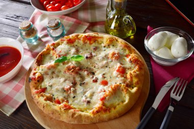 Yeni pişmiş Margherita pizzası ahşap bir kesim tahtasına dizilmiş. Yanında domates ve mozzarella peyniri var.