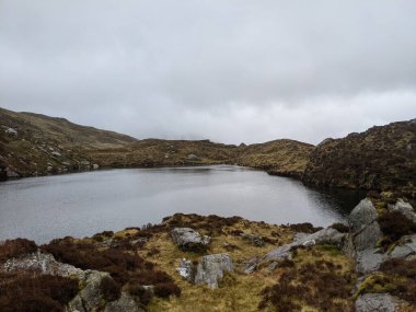 Kayalık kıyı ve bulutlu gökyüzü ile sakin bir dağ gölü. Sisin içinde Glyder Fawr 'a doğru yürüyüş, Snowdonia.