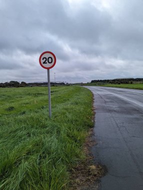 Çimenlerle ve bulutlu gökyüzüyle çevrili, düşük trafikli yolun yanında 20 mil hız uyarı levhası görünüyor..