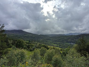 Yoğun ağaçları ve yuvarlanan tepeleri olan yemyeşil bir vadi üzerinde dramatik bulutlu bir gökyüzü. Manzara doğa görünümü.