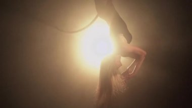 Young woman performs the acrobatic elements in the air hoop. Aerialist in on black background dark studio with backlight. For sports, acrobatic, circus school.