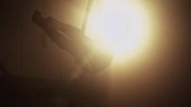 Young woman performs the acrobatic elements in the air hoop. Aerialist in on black background dark studio with backlight. For sports, acrobatic, circus school.