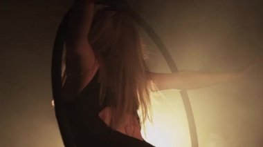 Young woman performs the acrobatic elements in the air hoop. Aerialist in on black background dark studio with backlight. For sports, acrobatic, circus school.