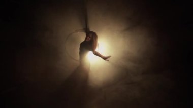 Young woman performs the acrobatic elements in the air hoop. Aerialist in on black background dark studio with backlight. For sports, acrobatic, circus school.