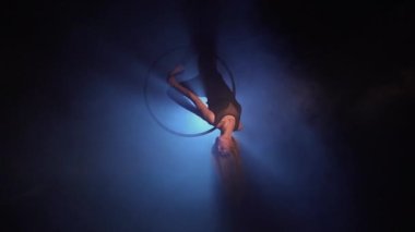 Young woman performs the acrobatic elements in the air hoop. Aerialist in on black background dark studio with backlight. For sports, acrobatic, circus school.