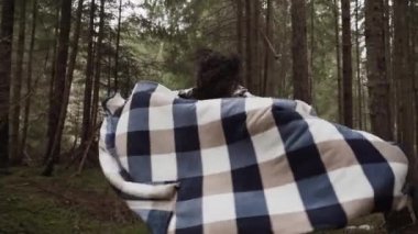 Sıcak bir battaniye ve şapkaya sarılı çekici bir kadın. Çam ormanında güneş ışığıyla poz veriyor. Dağ yolculuğu kavramı.