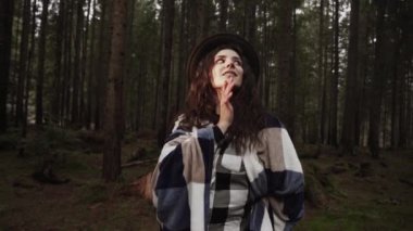Sıcak bir battaniye ve şapkaya sarılı çekici bir kadın. Çam ormanında güneş ışığıyla poz veriyor. Dağ yolculuğu kavramı.