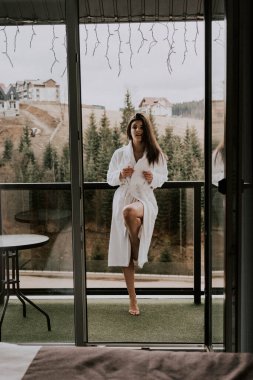 Beyaz bornozlu bir kadın kruvasan yer ve dağlardaki bir otelde kahve içer. Tatil konsepti.