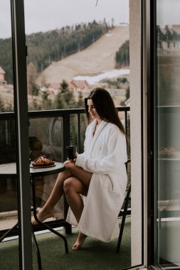Beyaz bornozlu bir kadın kruvasan yer ve dağlardaki bir otelde kahve içer. Tatil konsepti.