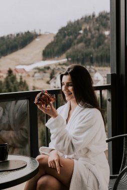 Beyaz bornozlu bir kadın kruvasan yer ve dağlardaki bir otelde kahve içer. Tatil konsepti.