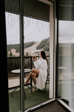 Beyaz bornozlu bir kadın kruvasan yer ve dağlardaki bir otelde kahve içer. Tatil konsepti.