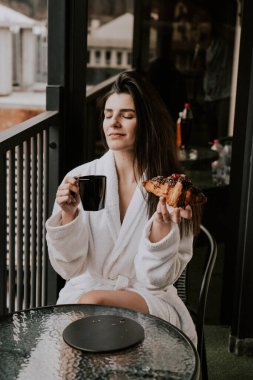 Beyaz bornozlu bir kadın kruvasan yer ve dağlardaki bir otelde kahve içer. Tatil konsepti.