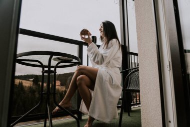 Beyaz bornozlu bir kadın kruvasan yer ve dağlardaki bir otelde kahve içer. Tatil konsepti.