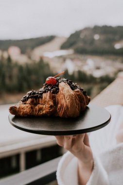 Beyaz bornozlu bir kadın kruvasan yer ve dağlardaki bir otelde kahve içer. Tatil konsepti.