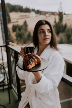 Beyaz bornozlu bir kadın kruvasan yer ve dağlardaki bir otelde kahve içer. Tatil konsepti.