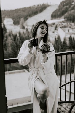 Beyaz bornozlu bir kadın kruvasan yer ve dağlardaki bir otelde kahve içer. Tatil konsepti.