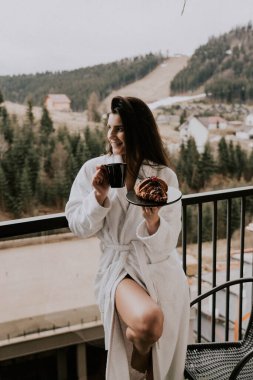 Beyaz bornozlu bir kadın kruvasan yer ve dağlardaki bir otelde kahve içer. Tatil konsepti.