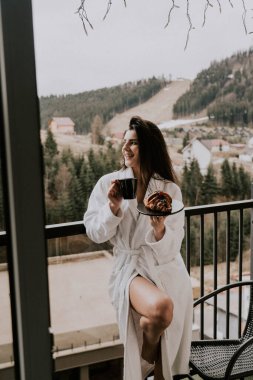 Beyaz bornozlu bir kadın kruvasan yer ve dağlardaki bir otelde kahve içer. Tatil konsepti.