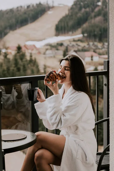 Beyaz bornozlu bir kadın kruvasan yer ve dağlardaki bir otelde kahve içer. Tatil konsepti.
