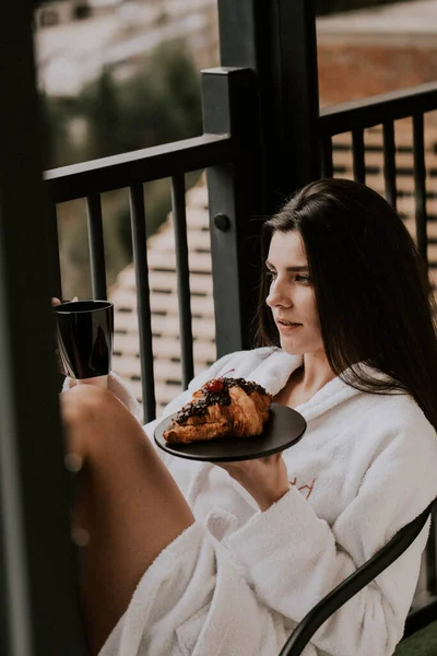 Beyaz bornozlu bir kadın kruvasan yer ve dağlardaki bir otelde kahve içer. Tatil konsepti.