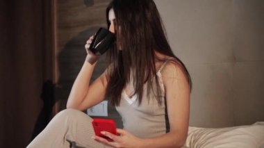 Akıllı telefon kullanan ve sabahın köründe yatakta sıcak kahve içen mutlu bir kadın. Akıllı telefon ve elinde kahveyle yatakta genç bir kadın. Cep telefonu ve internet kullanan mutlu bir kadın.