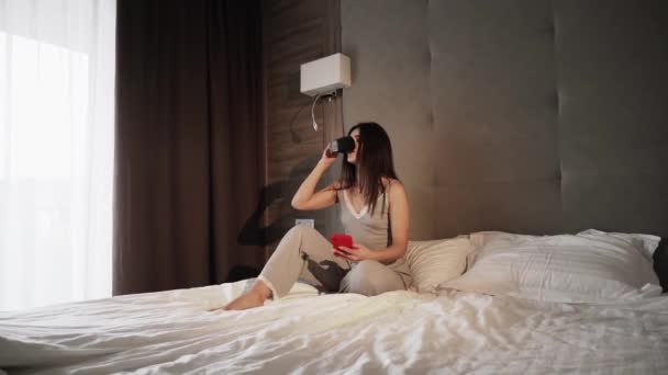 快乐的微笑的女人早上在床上用智能手机泡热咖啡 年轻的女人手里拿着智能手机和一杯咖啡躺在床上 快乐的女性使用手机和互联网 — 图库视频影像