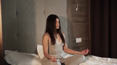 Pijamalı genç bir kadın büyük bir yatakta lotus pozu vererek meditasyon yapıyor. 