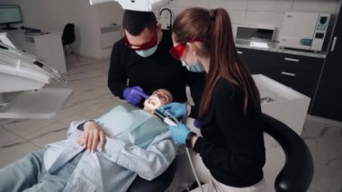 Diş kliniğinde koruyucu maskeli iki doktor hasta kıza hizmet ediyor. İş Dişçiliği. Sağlık ve Tıp Konsepti.